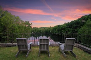 Trois chaises sont placées face à un lac paisible, avec une forêt en arrière-plan et un ciel aux couleurs vives.