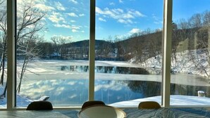 Ein Zimmer mit großem Fenster und Blick auf einen zugefrorenen See sowie umliegende Bäume.