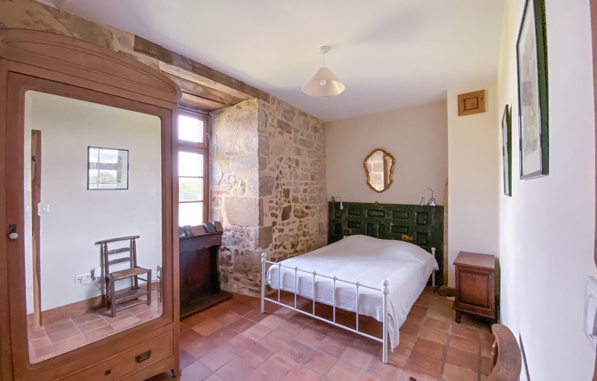 Une chambre avec un mur en pierre, une armoire en bois, un lit recouvert d’une courtepointe blanche, une tête de lit verte et une petite table de chevet.
