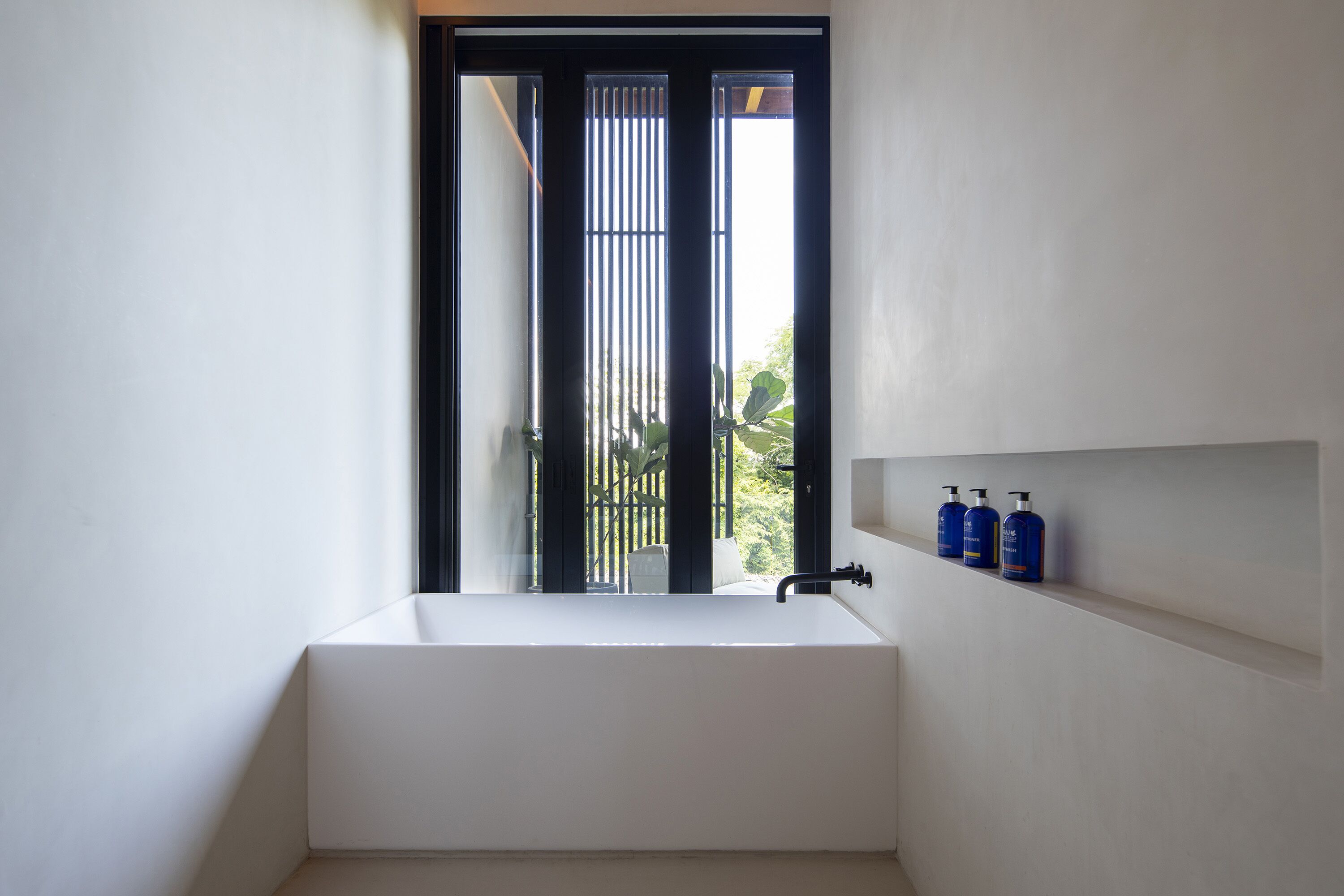 A modern bathroom with a large white bathtub, a window with vertical blinds, and a shelf with three blue bottles.