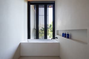 A modern bathroom with a large white bathtub, a window with vertical blinds, and a shelf with three blue bottles. - Boho Villa - Brand New 4 Master BD (Cóbano)
