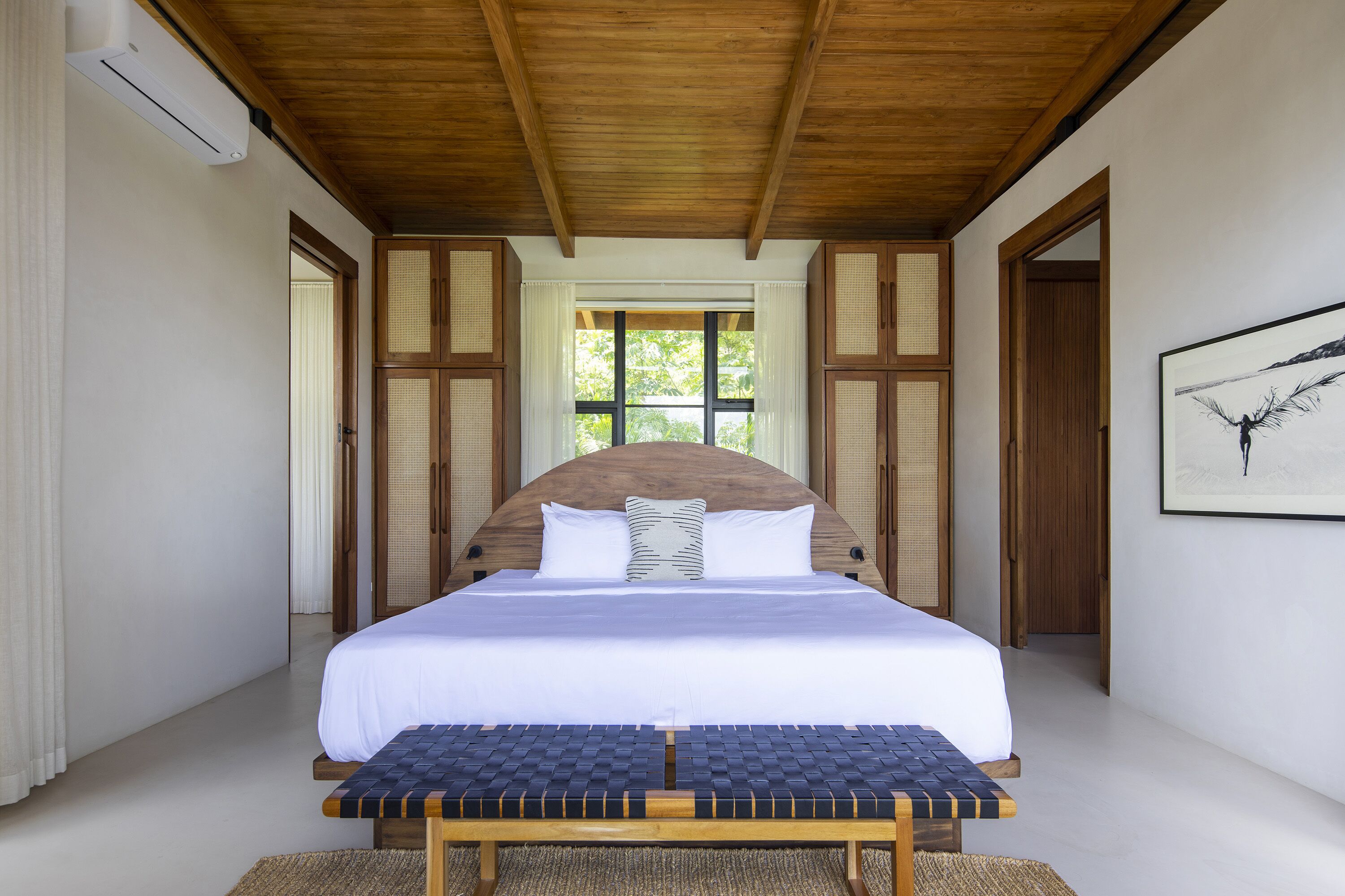A bedroom with a large bed, a bench at the foot of the bed, wooden ceiling beams, and a window with white curtains.