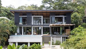 Una casa moderna de dos pisos con un amplio balcón de cristal, rodeada de exuberante follaje.