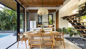 Un comedor con mesa y sillas de madera, una lámpara colgante y vistas de la cocina y la piscina exterior.