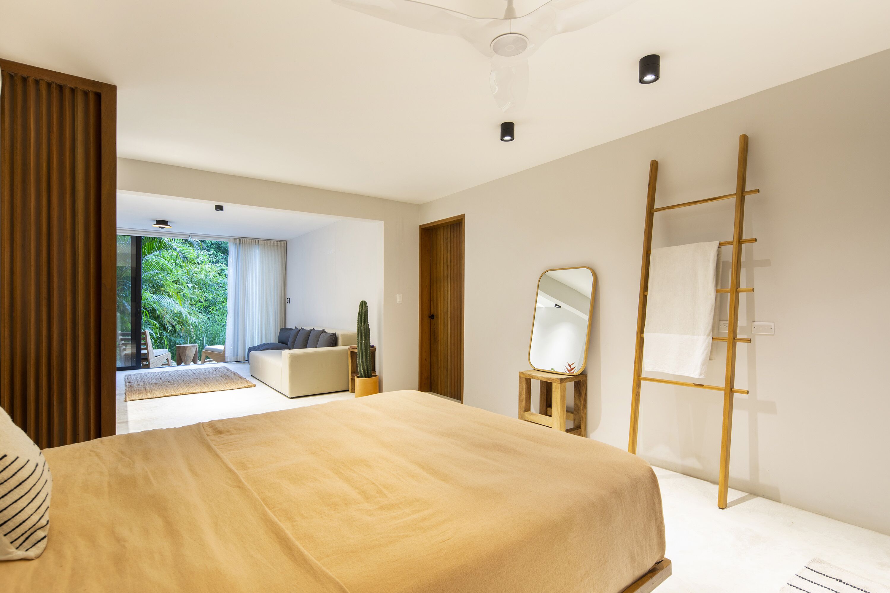 A modern bedroom with a large bed, a wooden ladder, a mirror, and a view of a balcony with outdoor furniture.