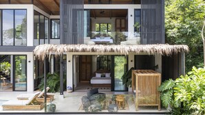Una casa moderna de dos pisos con techo de paja, patio, ventanas amplias y un dormitorio visible con cama y zona de estar.