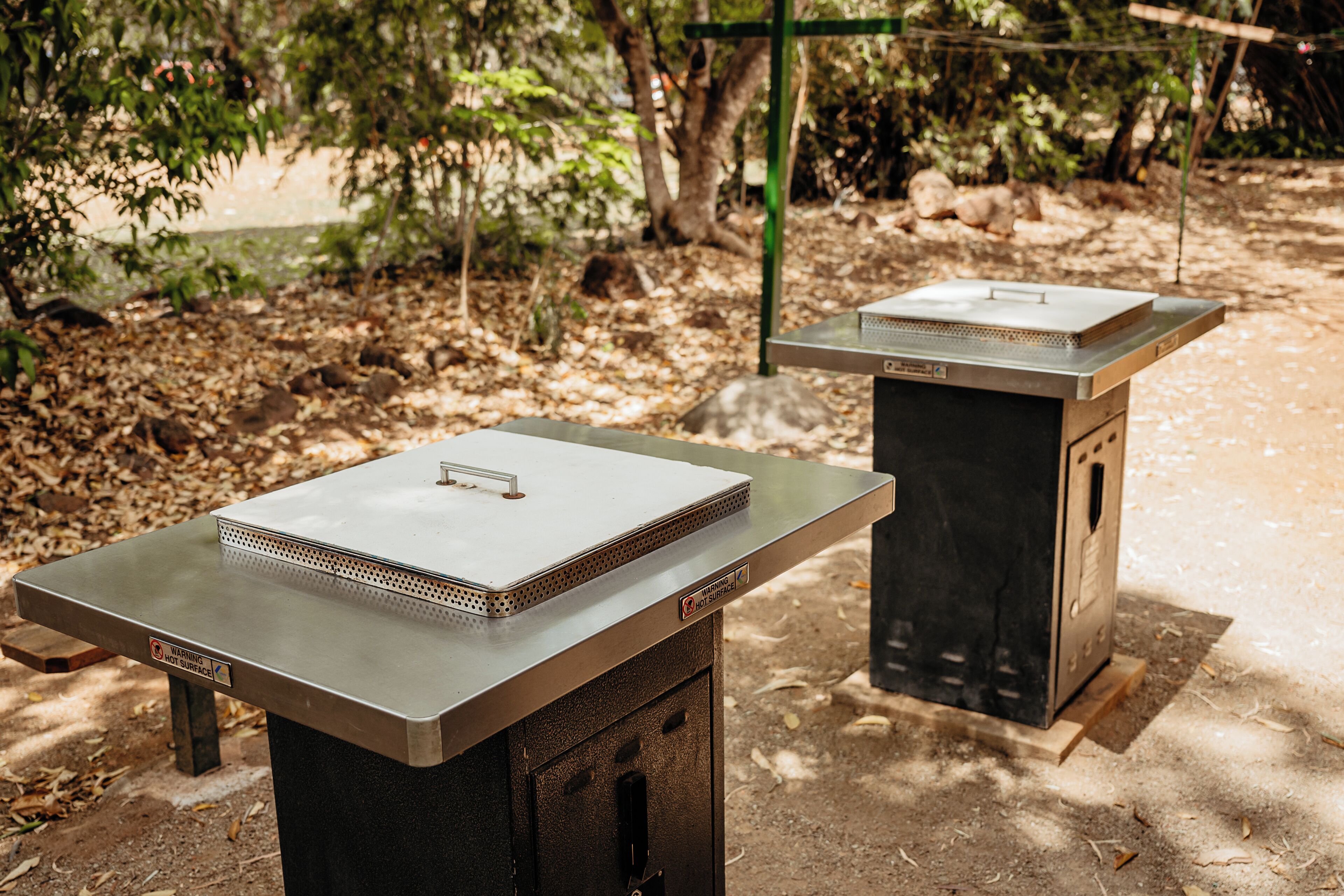 Two outdoor barbecue grills with metal tops and ventilation grates, set on a concrete surface with a backdrop of trees and a dirt path.
