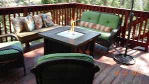 An outdoor deck with a fire pit table, green cushioned seating, and a wicker chair. - 3 BR+loft, fam. rm., 2 bth, lg deck, close to major ski areas, trails, history. (Soda Springs)