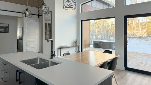 A modern kitchen with a large island, pendant lighting, and a view of a snowy outdoor deck. - Modern Riding Mountain Retreat (Onanole)