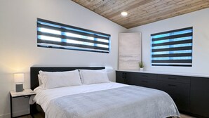 A modern bedroom with a wooden ceiling, a bed with white bedding, a nightstand with a lamp, and two wall-mounted windows with blinds. - Modern Riding Mountain Retreat (Onanole)