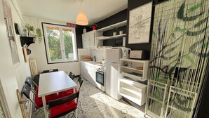 Une cuisine compacte avec des meubles blancs, une table à manger avec des chaises, un luminaire suspendu et une fenêtre donnant sur un espace verdoyant.