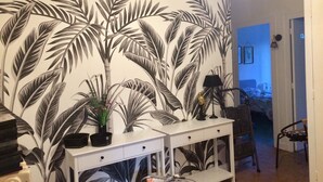 Une pièce décorée d’une grande fresque murale représentant des feuilles de palmier, avec une console blanche dotée de tiroirs, une lampe et une chaise.