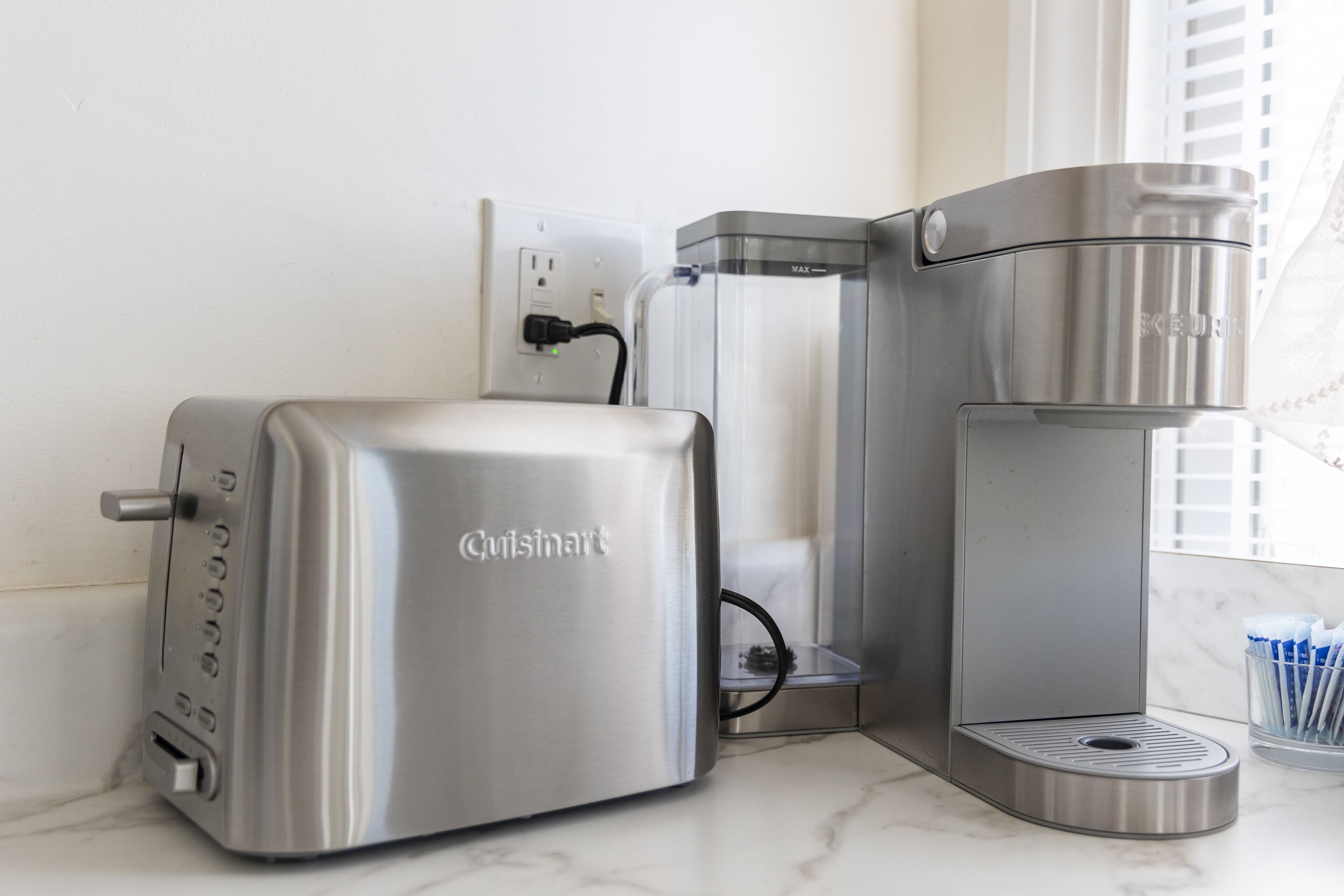 A kitchen with a stainless steel Cuisinart toaster and coffee maker, a power outlet, and a window with blinds.