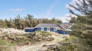 Une maison avec une allée en gravier menant à une porte en bois, entourée d’une végétation naturelle et d’arbres.