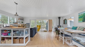 Une cuisine spacieuse avec un plancher en bois, un coin repas avec une table et des chaises, un fauteuil jaune et une vue sur l’extérieur grâce à de grandes fenêtres.
