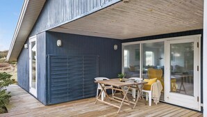 Un espace extérieur abrité, doté d’une terrasse en bois, d’une table avec des chaises et d’une porte-fenêtre coulissante donnant sur un espace intérieur.