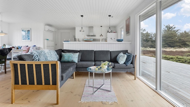 Un salon moderne avec un canapé gris, une chaise en bois et une table basse en verre. On aperçoit, au fond, une cuisine avec des meubles blancs et une grande fenêtre.