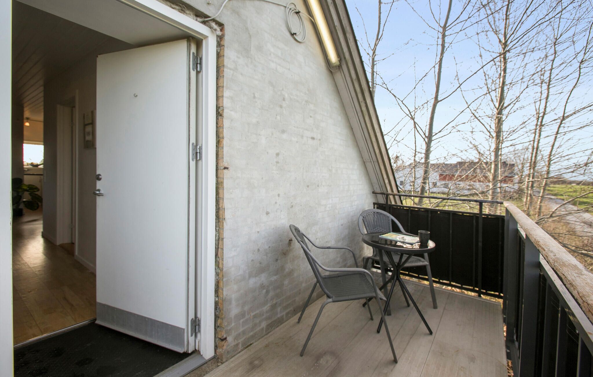 Een balkon met een tafel en stoelen, een deur die naar een binnenruimte leidt, en een uitzicht op bomen en gebouwen op de achtergrond.