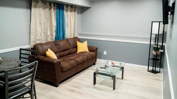 A living room with a brown sofa, two yellow cushions, a glass coffee table, a dining table with chairs, and a TV mounted on the wall. - JC Entire Apartment remodeled, close to Airport and Ferry (Coram)