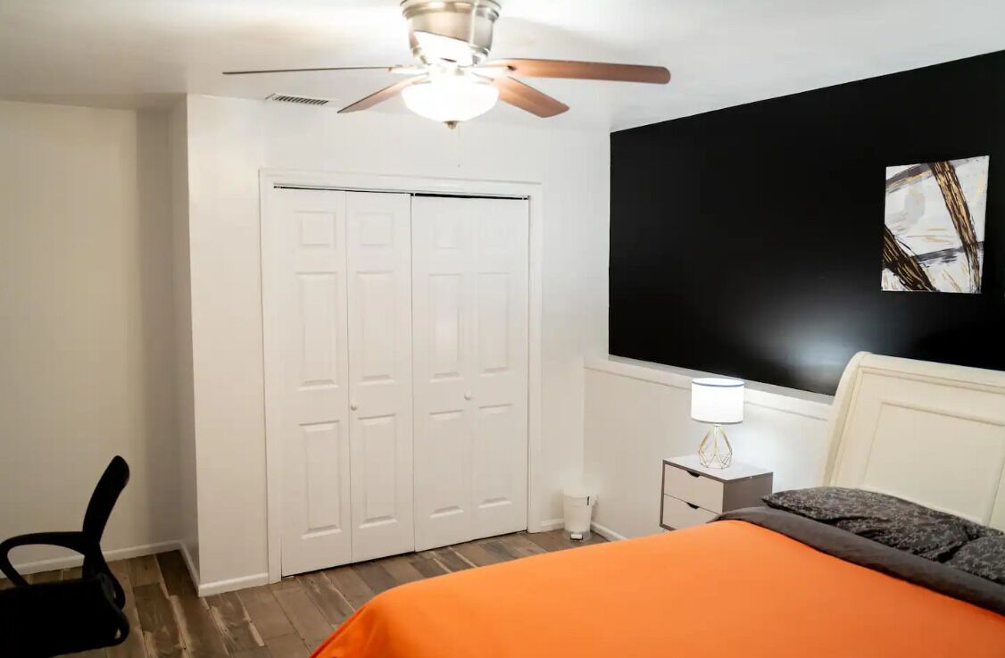 Un dormitorio con ventilador de techo, cama con colcha naranja, mesita de noche con lámpara y un cuadro abstracto en blanco y negro en la pared.