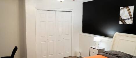 Un dormitorio con ventilador de techo, cama con colcha naranja, mesita de noche con lámpara y un cuadro abstracto en blanco y negro en la pared.