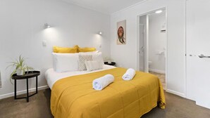 Ein modernes Hotelzimmer mit Bett, einem kleinen Beistelltisch, einer Topfpflanze und einer Tür zu einem Badezimmer.