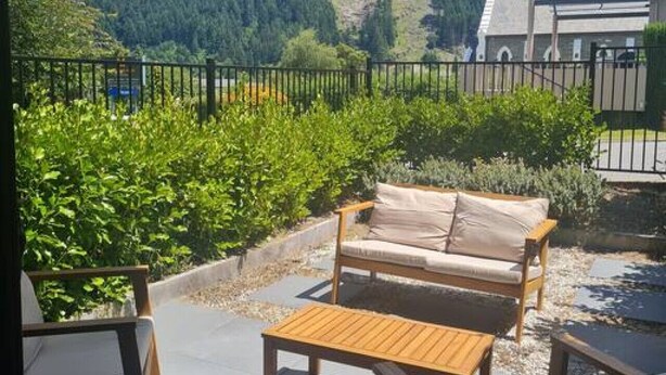 Eine Terrasse mit einer Holzbank, einem Sofa und einem Tisch. Im Hintergrund sind ein Gebirge und ein Gebäude zu sehen.
