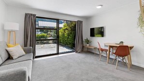 Ein modernes Wohnzimmer mit einem grauen Sofa, einem Esstisch mit zwei Stühlen, einem flachbildfernseher an der Wand und einer Glastür, die auf einen Balkon mit Grünpflanzen führt.