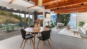 Ein modernes Interieur mit Holzbalkendecke, einem Essbereich mit rundem Tisch und Stühlen, einer Couch und Blick auf einen See.