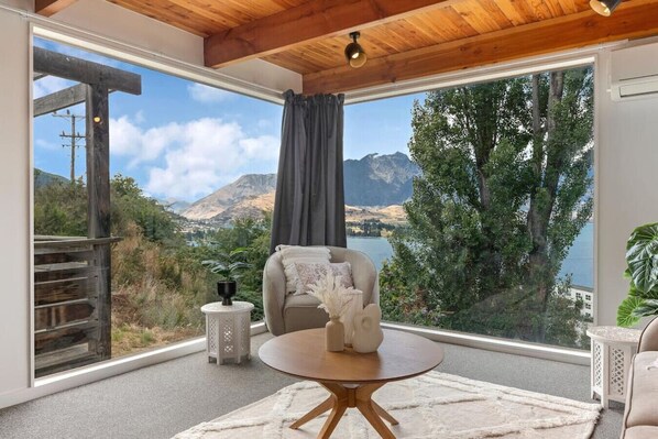 A room with a large window offering a mountain and lake view, a wooden ceiling, and a round table with a vase.