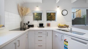 Una cocina moderna con armarios blancos, una placa de inducción negra y un lavamanos de acero inoxidable. Hay dos ventanas que aportan luz natural, y un reloj en la pared.