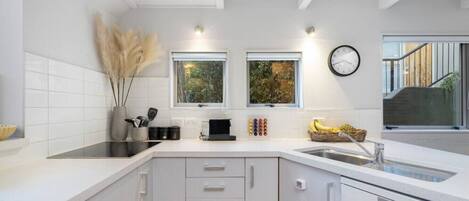 A modern kitchen with white cabinetry, a black induction cooktop, and a stainless steel sink. There are two windows providing natural light, and a clock on the wall.