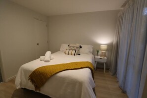 A neatly made bed with a white comforter and a mustard-colored throw blanket. There is a bedside table with a lamp and a small vase with flowers. The room has light-colored walls and a wooden floor. - 5 minute easy walk to Queenstown, warm and spacious. (Queenstown)