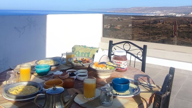 Et spisebord med forskelligt mad og drikke, placeret på en balkon med udsigt til havet og det fjerne landskab.