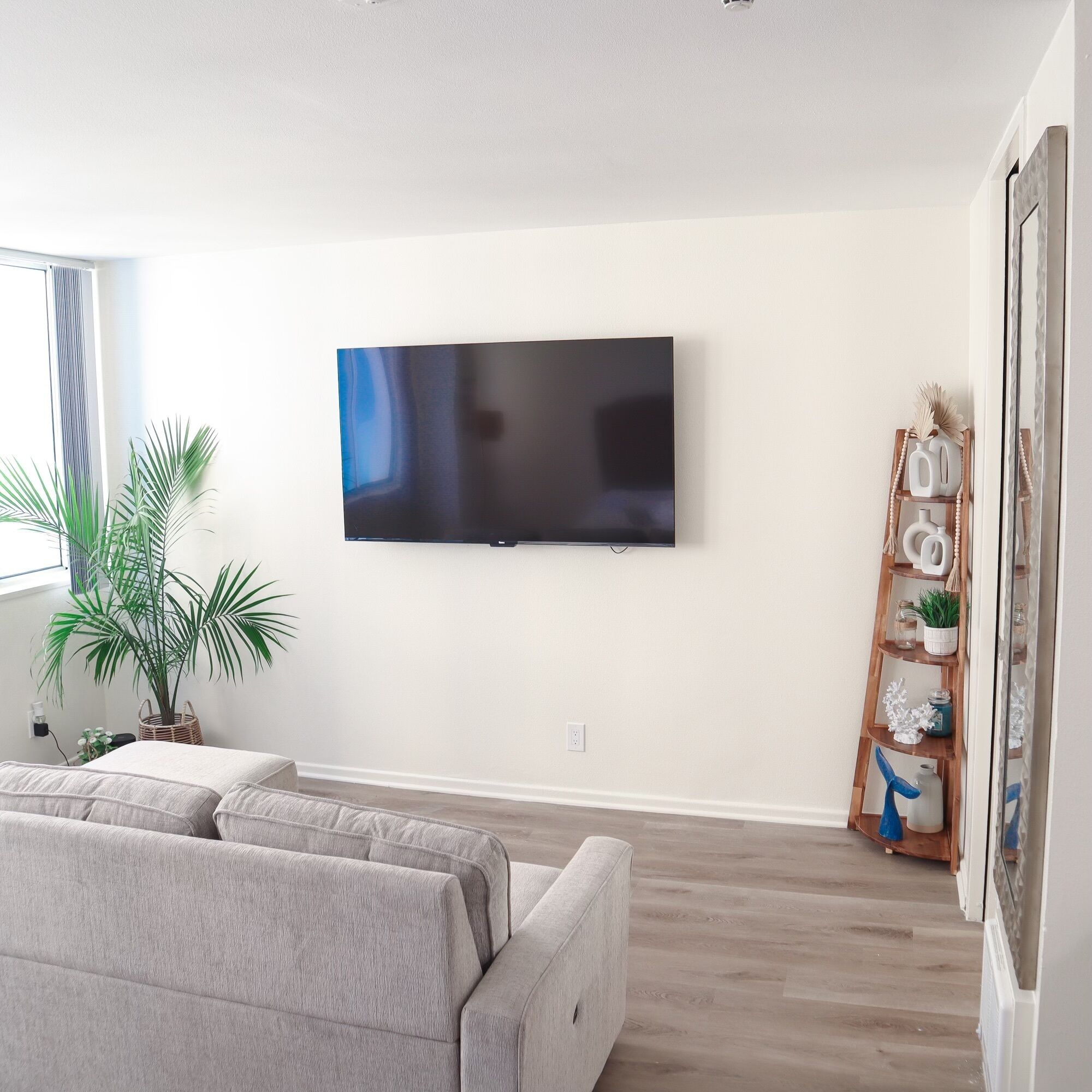 Ein Wohnzimmer mit flachbildfernseher an der Wand, einem grauen Sofa, Holzboden und einem dekorativen Regal mit verschiedenen Gegenständen.