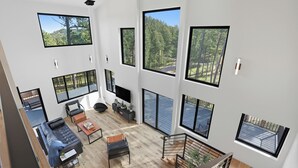A spacious living area with a large sofa, coffee table, and TV, featuring multiple large windows offering views of a forested area. - The Falcon- Breathtaking Mountain Views (Rapid City)