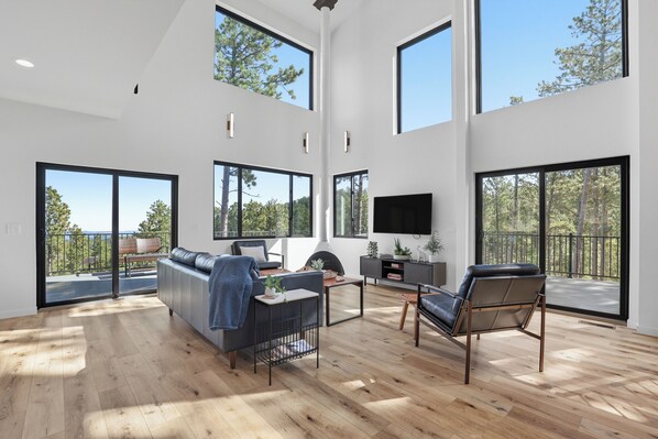 A spacious living room with a large sofa, a coffee table, and a fireplace. There are large windows providing natural light and views of the outdoors. - The Falcon- Breathtaking Mountain Views (Rapid City)
