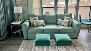 Ein Wohnzimmer mit einem grünen Sofa, passenden Hocker, einem Beistelltisch mit Lampe und Blick auf eine verschneite Landschaft durch große Fenster.
