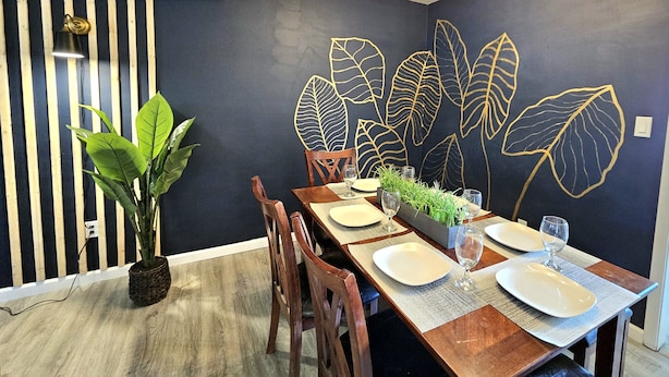 Un coin repas avec un mur bleu foncé, une table à manger en bois dressée avec des assiettes et des verres, et une plante en pot.