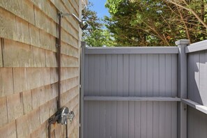 A grey wooden fence with a metal gate, a metal pipe, and a tree in the background.