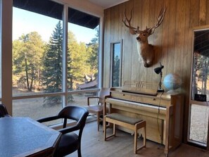 A room with a piano, a deer head mounted on the wall, and a view of trees through large windows. - Beautiful Hall Creek Estate (Greer)