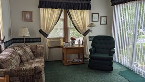 A living room with a patterned sofa, a green armchair, a small wooden side table with a vase and magazines, and a window with curtains. - Park model unit #2 for rent by Silver Lake NY  (Perry)