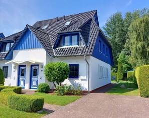 Una casa de dos pisos con fachada azul y blanca, techo de tejas y un jardín cuidado con setos y un árbol pequeño.
