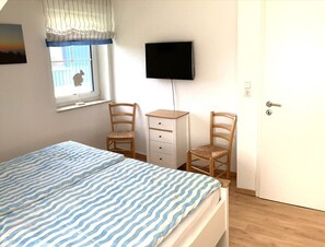 Un dormitorio con cama, dos sillas, escritorio, televisor y ventana con cortinas.