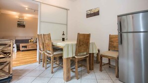 Een eetgedeelte met een houten tafel en stoelen, een koelkast, en uitzicht op een woonkamer met een bank en een schilderij aan de muur.