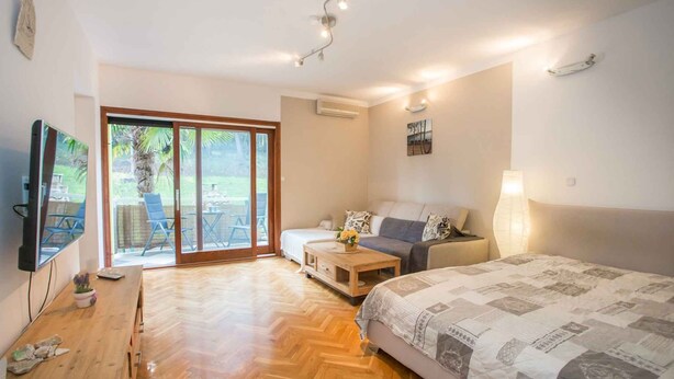 Een kamer met een bed, een slaapbank, een televisie, een houten tafel en een glazen schuifdeur die leidt naar een buitenruimte.