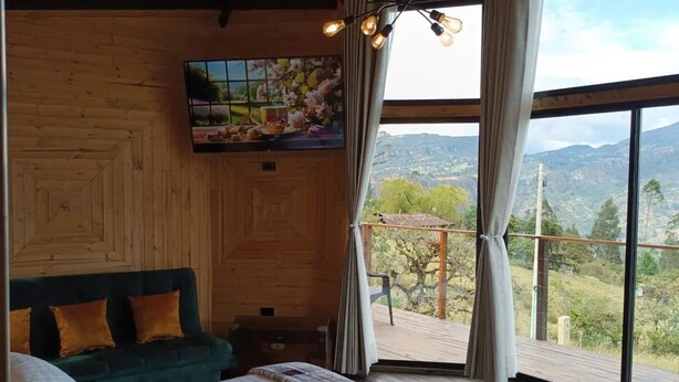 A bedroom with a bed, a green sofa, a TV on the wall, and a large window with a view of the outdoors. - Glamping Caelum, Refugio al Aire Libre. Macheta Cundinamarca (Machetá)