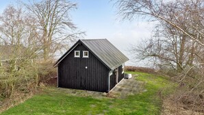 A small, dark wooden building with a sloped roof and two windows, surrounded by grassy fields and leafless trees.