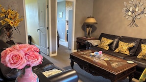 A living room with a dark brown coffee table, a black leather sofa with patterned cushions, a pink vase with flowers, and a wall-mounted lamp. - Quiet cozy room no stairs in a peaceful house, safe community, full kitchen (Langley Township)