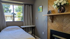 A bedroom with a bed, a bedside table, a window with curtains, a wall-mounted shelf with flowers, and a fireplace. - Quiet cozy room no stairs in a peaceful house, safe community, full kitchen (Langley Township)